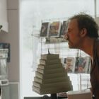 Zwei Menschen in einem Laden interagieren ernsthaft miteinander. Links ist eine Frau, rechts ein Mann mit einem Papiertaschentuch in der Nase. Im Hintergrund sind Regale mit DVDs zu sehen.