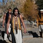 Drei Männer in traditioneller Kleidung mit Federschmuck und Bemalung im Gesicht stehen in einer felsigen, bewachsenen Landschaft.