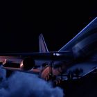 Ein großes Flugzeug fliegt durch den Nachthimmel, aus einem Triebwerk kommt Feuer.