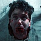 Es ist keine Schrift im Bild. Ein Kopf steckt durch ein Loch in einer Tür, das Gesicht hat Blutspuren.