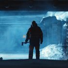 Keine sichtbaren Texte. Eine Person mit Axt steht im Schnee, umgeben von Blockhütten, in kaltem, blauen Licht.