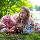 Zwei Mädchen liegen auf einer Decke im Grünen am Wasser, eins schläft, eins schaut nachdenklich. Im Hintergrund steht jemand.