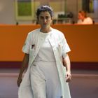 Eine Frau in einem weißen Kittel geht durch einen Krankenhausflur. Im Hintergrund ist eine orangefarbene Theke.