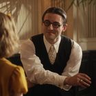 Ein Mann mit Brille und Anzug sitzt an einem Klavier und spricht mit einer Person in gelber Kleidung.