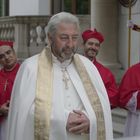 Männer in roter Kirchenkleidung und ein Mann in weißen Gewändern mit goldenem Schal, im Hintergrund ein Soldat.