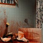 Ein Badezimmer mit hellblauen Fliesen, Blut spritzt an Wänden und Boden. Eine umgefallene Figur, zerschlagenes Keramikobjekt.