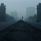 In nebliger Stadtlandschaft stehen monumentale Torbögen, wenige Autos auf leerer Straße, düstere Atmosphäre.