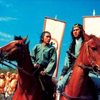 Zwei Reiter in traditionellen asiatischen Gewändern, himmelblauer Hintergrund, gefolgt von Männern in Orange.