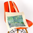 Eine Stofftasche mit einer Impressionismus-Malerei von einer Brücke liegt auf einem knallroten Stuhl.