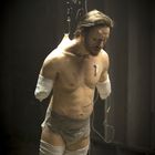 Ein Mann ohne Arme und Beine hängt an Schläuchen in dunkler Umgebung. Sein Gesicht wirkt leidend.