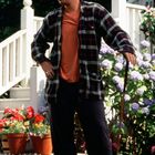 Ein Mann in kariertem Hemd und Baseballkappe steht mit einem Golfschläger vor bunten Blumen und einer weißen Treppe.