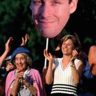 Zwei Frauen klatschen glücklich; eine Frau trägt einen blauen Hut, die andere einen weißen Pullunder. Im Hintergrund ein großes Gesicht.