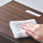 Keine sichtbaren Texte. Eine Hand wischt Wasser von einem Holztisch mit einem weißen Tuch.