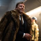 Ein Mann im Anzug mit einem großen, pelzbesetzten Mantel, umgeben von Personen in Militäruniformen mit blauen Baretten.