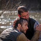 Zwei Männer in nasser Kleidung umarmen sich am Fluss. Einer wirkt erschöpft, der andere tröstet ihn.