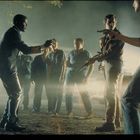 Eine Gruppe von Männern steht im Freien. Einer hält eine Pistole, ein anderer beschwichtigt mit erhobenen Händen. Es ist Nacht.