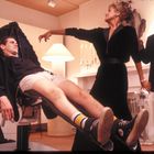 Ein Mann in Unterwäsche liegt in einem Stuhl, während eine Frau und ein Mann im Anzug daneben stehen.