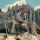 Ich kann die Texte im Bild nicht lesen. Eine Person wandert, umgeben von bizarren Felsformationen und Seilen, bei sonnigem Wetter.