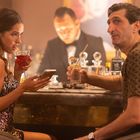 Ein Mann und eine Frau sitzen in einer eleganten Bar, trinken Cocktails. Sie sieht auf ihr Handy, während er in die Ferne schaut.