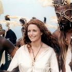 Eine lächelnde Frau in weißer Bluse steht zwischen Männern in traditionellen afrikanischen Masken und Kostümen.