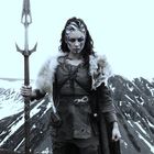 Eine Kriegerin in Lederrüstung, Pelz und Kriegsbemalung steht vor einer schneebedeckten Berglandschaft, hält einen Speer.