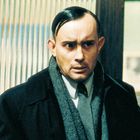 Ein besorgter Mann in Anzug und Schal steht vor vertikalen Streifen.