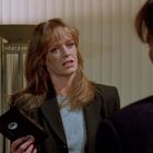 Eine Frau mit langen Haaren hält eine VHS-Kassette und spricht mit einem Mann in einem Raum mit vertikalen Jalousien.