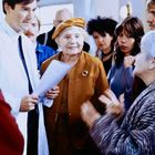 Ein Arzt hält Dokumente. Ältere Dame in braunem Outfit spricht, umgeben von mehreren Personen im Gespräch vertieft.