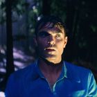 Eine Person mit nassem Haar in einem dunklen Wald, ausdrucksvoll beleuchtet.