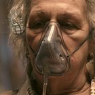 Eine ältere Person mit grauen Haaren trägt eine Sauerstoffmaske. Die Stimmung ist ernst und konzentriert.