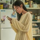 Eine Frau in einem gelben Oberteil schaut auf ihr Smartphone. Im Hintergrund sind eine Küche und ein Kühlschrank stehen.