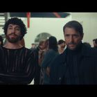Zwei Männer, einer mit Locken, der andere mit Bart, stehen inmitten einer geselligen Menschenmenge in elegantem Umfeld.