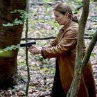 Keine Texte sichtbar. Eine Frau im braunen Mantel zielt mit einer Pistole im Wald. Bäume und Blätter umgeben sie.