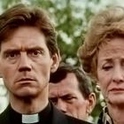 Ein ernster Mann in Priesterkleidung und eine besorgte Frau stehen dicht nebeneinander, im Hintergrund unscharfe Bäume.