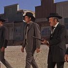 Viele Schilder an Gebäuden, vier Männer in Anzügen und Hüten laufen in einer Wüstenstadt.