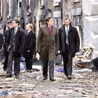 Mehrere Männer in Anzügen gehen durch eine heruntergekommene städtische Gegend voller Trümmer.