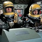 Zwei Personen in Raumanzügen mit gelben Helmen sitzen in einem Cockpit voller Instrumente und Bildschirme.