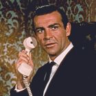 Ein Mann im Anzug hält einen Telefonhörer, vor einem gemusterten, luxuriösen Hintergrund.