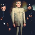 Zwei Männer in schwarzen Uniformen neben einem Mann in beigem Anzug, alle schauen ernst. Die Uniformen zeigen ein Blitzsymbol.