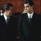 Zwei Männer in Anzügen stehen nebeneinander und sprechen, im Hintergrund unscharfe Personen in einem Raum mit Holzverkleidung.