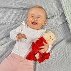 Ein lächelndes Baby in einem weißen Shirt mit Herzmustern hält ein Kuscheltier in einem roten Outfit und liegt auf einer grauen Decke.