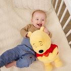 Der gelbe Teddy trägt ein rotes Shirt mit dem Wort "Pooh". Ein lächelndes Baby liegt daneben im Kinderbett.