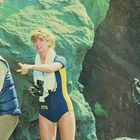 Eine Frau im blauen Taucheranzug zeigt mit der Hand. Zwei Männer neben ihr, einer mit Gewehr, einer mit Harpune. Felsiger Hintergrund.