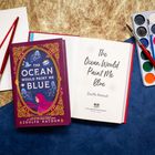 "THE OCEAN WOULD PAINT ME BLUE - Zoulfa Katouh" steht auf Buch und Seite, umgeben von Pinseln und Farben.