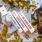 Drei Bücher: "The Grandest Game", "Glorious Rivals", "The Gilded Blade". Umgeben von Spielkarten und goldenen Blättern.