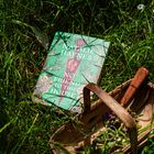 „NATALIE HAYNES“, „NO FRIEND TO THIS HOUSE“. Buch liegt im Gras neben einem Weidenkorb mit Blumen und Gartenwerkzeug.