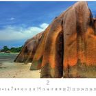 "La Digue, Seychellen" unten links. Große orange-braune Granitfelsen an einem Strand, blauer Himmel und Meer im Hintergrund.