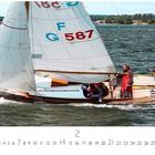 G-587, 2. Segelboot auf leicht welligem Wasser. Zwei Personen in roter Kleidung. Im Hintergrund eine Küstenlandschaft.