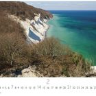 Nationalpark Møen. Küste mit weißen Klippen, blau-grünes Meer, bewaldete Hügel und kahler Baumbestand im Vordergrund.