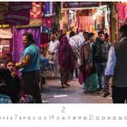 Aufschrift: "Dehli, Basar". Zahl "2" darunter. Menschen in bunten Kleidern auf einem belebten Markt, umgeben von Stoffen.